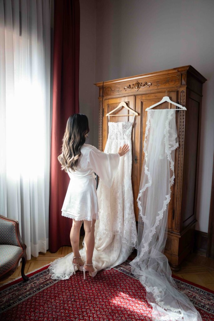 A woman in a white gown touches a wedding dress and veil hanging on a wooden wardrobe in a sunlit room – perfect for wedding photographers capturing unforgettable moments of an abroad wedding.