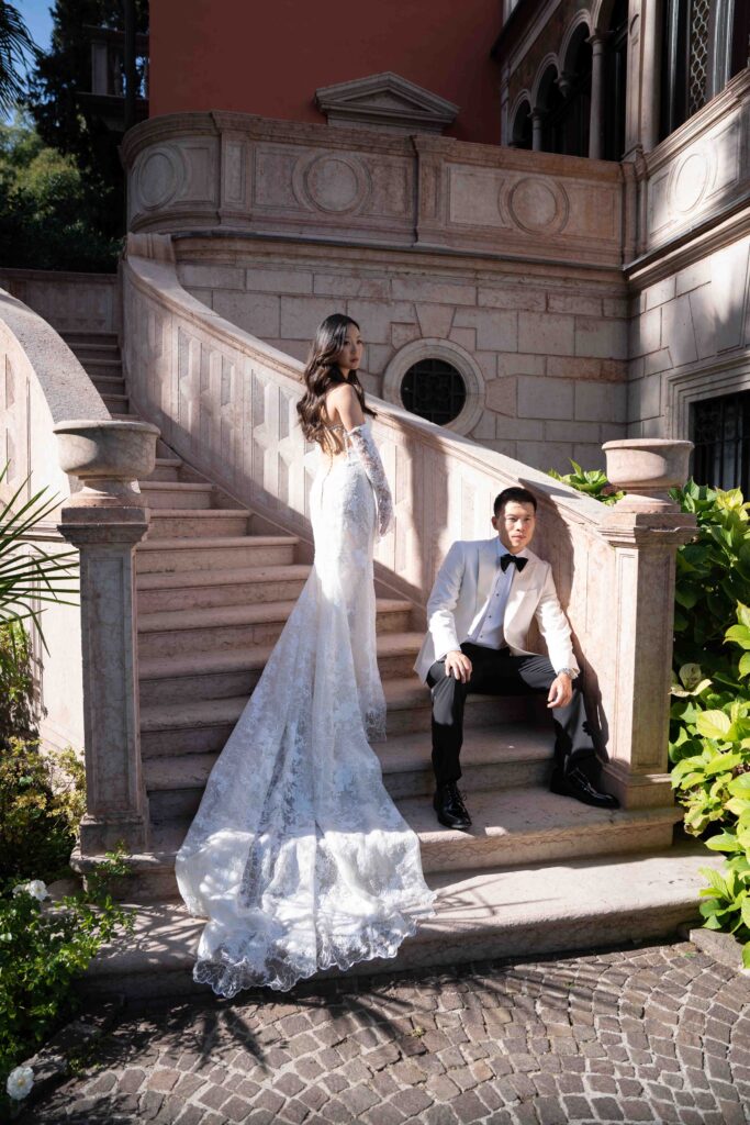 A bride in a white dress stands on a staircase while the groom in a white tuxedo jacket sits below her, capturing a timeless moment of overseas wedding photography.