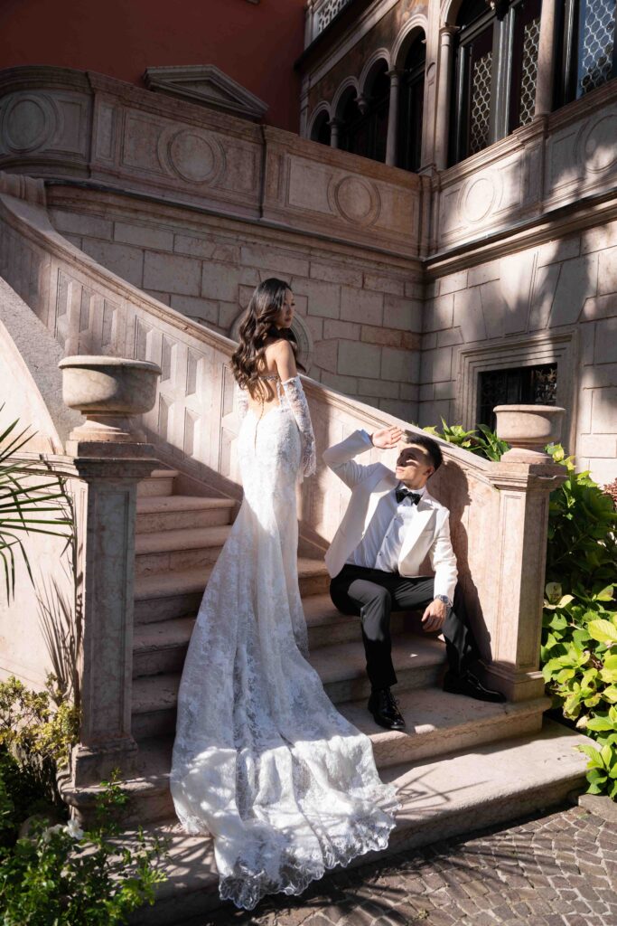 Heiraten am Gardasee: Locations, Kosten und ehrliche Erfahrungen aus über 15 Hochzeiten 41 Eine Braut in einem langen Spitzenkleid steht auf einer Treppe, während der Bräutigam im Smoking unten sitzt und sie bewundert - ein perfekter Moment, eingefangen von einer Hochzeitsfotografin, ideal für Hochzeiten am Urlaubsort.