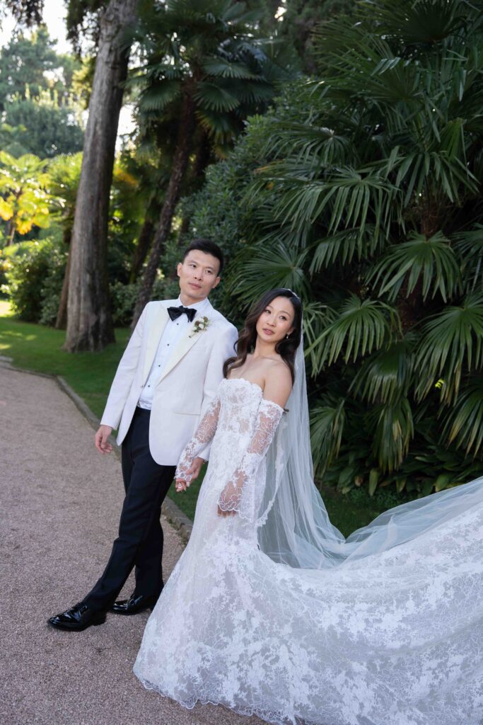 Die Braut in einem schulterfreien Spitzenkleid und der Bräutigam in einem weißen Smoking posieren im Freien inmitten von üppigem Grün - perfekt für Hochzeiten am Urlaubsort und unvergessliche Hochzeitsfotografien.