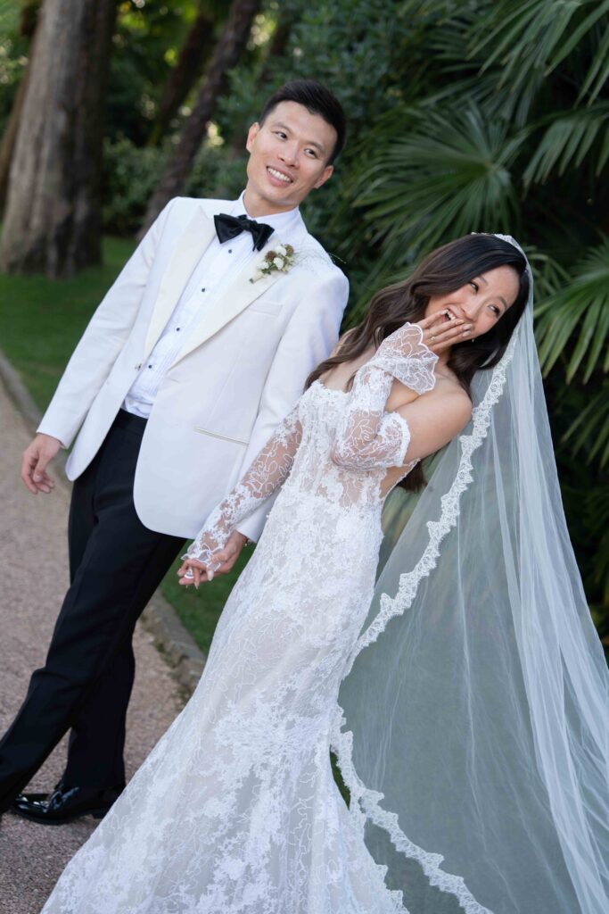 Braut im Spitzenkleid und Bräutigam im weißen Smoking lächelnd und händchenhaltend im Freien, umgeben von Grün - ein perfekter Moment, eingefangen von einer erfahrenen Hochzeitsfotografin. Hochzeitsfotografie im Ausland für unvergessliche Erinnerungen.