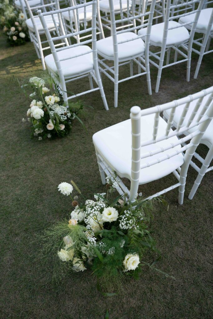 Weiße Stühle im Freien mit Blumenarrangements auf dem Gras daneben, perfekt für eine romantische Auslandshochzeit oder eine Hochzeit am Reiseziel.