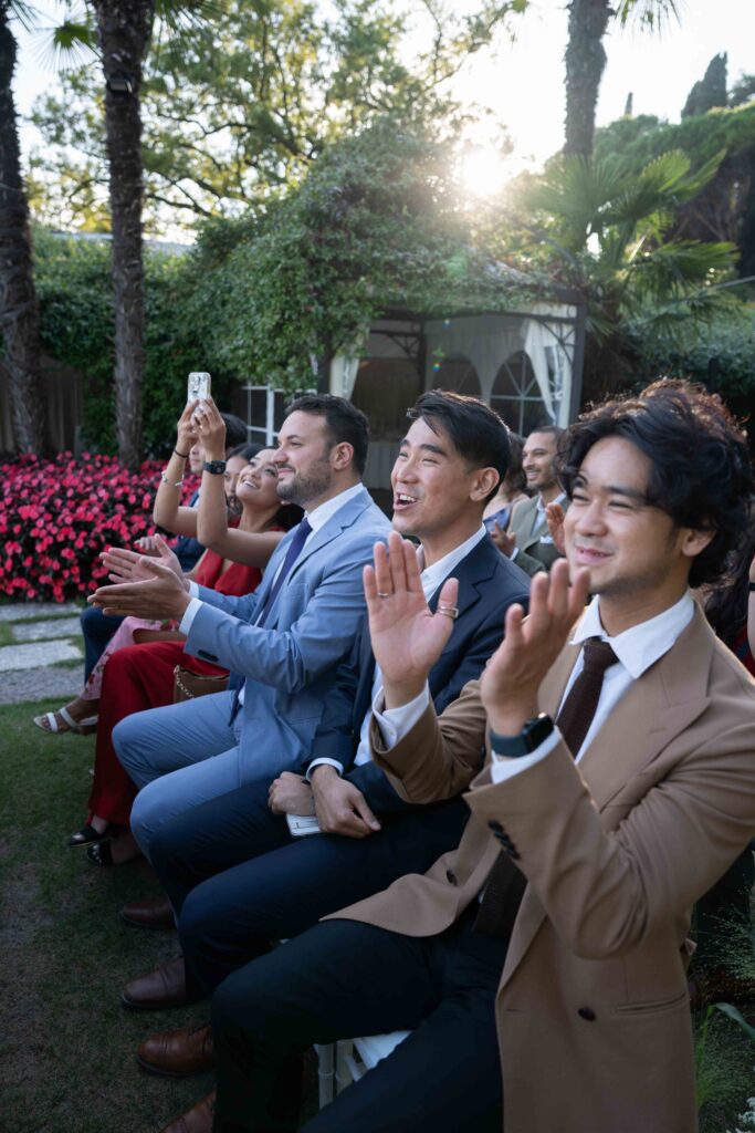Editorial Wedding Photography: The Trend for Stylish Wedding Photos Guests in formal attire are seated outdoors, smiling and applauding at a beautiful destination wedding, perfectly captured by wedding photographers who specialise in overseas wedding photography.