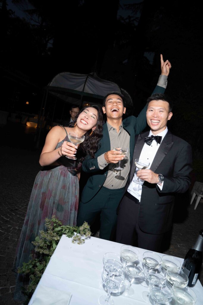 Editorial Wedding Photography: The Trend for Stylish Wedding Photos 5 Three formally dressed people smile and toast with a drink at an outdoor evening event. They capture the joy and elegance often seen at destination weddings – the perfect inspiration for overseas wedding photography.