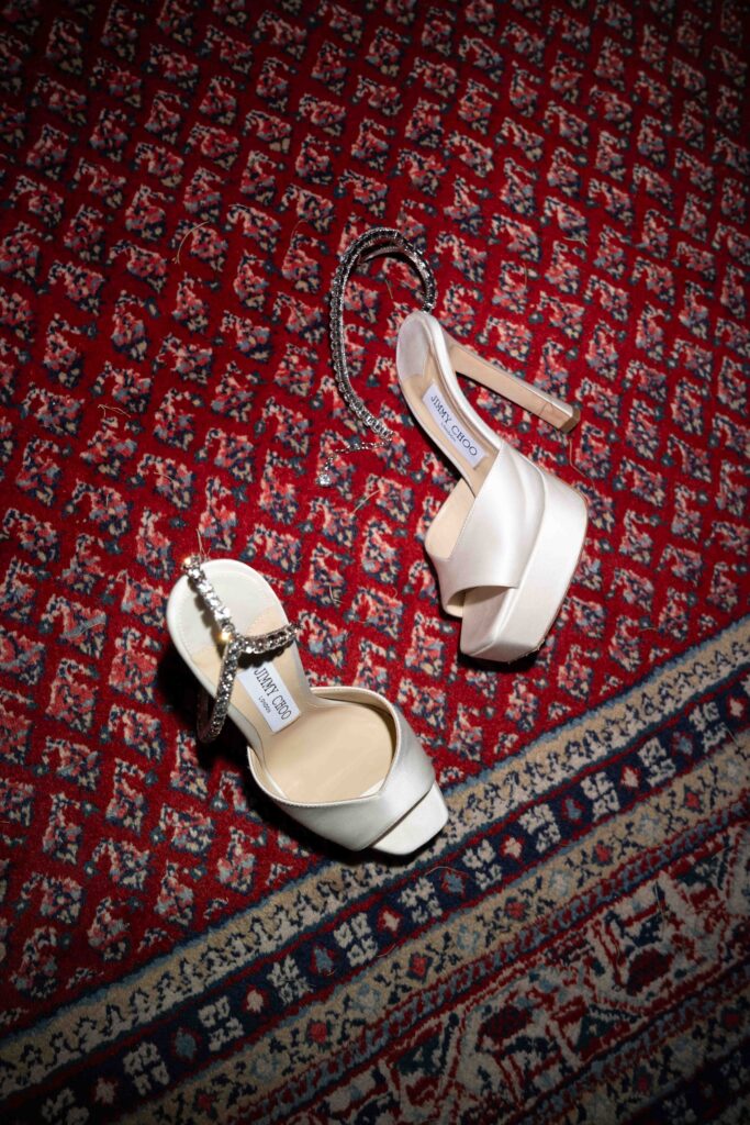 Two elegant stiletto heels with ornate straps rest on a red and white patterned rug – the perfect inspiration for any wedding photographer and lovers of stylish wedding photography.
