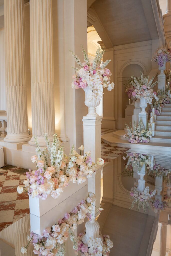 Elegante Blumenarrangements aus pastellfarbenen Blüten schmücken einen prächtigen Saal mit Säulen und Marmorboden und schaffen so das Ambiente eines automatisch zusammengestellten Meisterwerks – wie ein automatisch gespeicherter Entwurf, der anmutig zum Leben erweckt wurde.