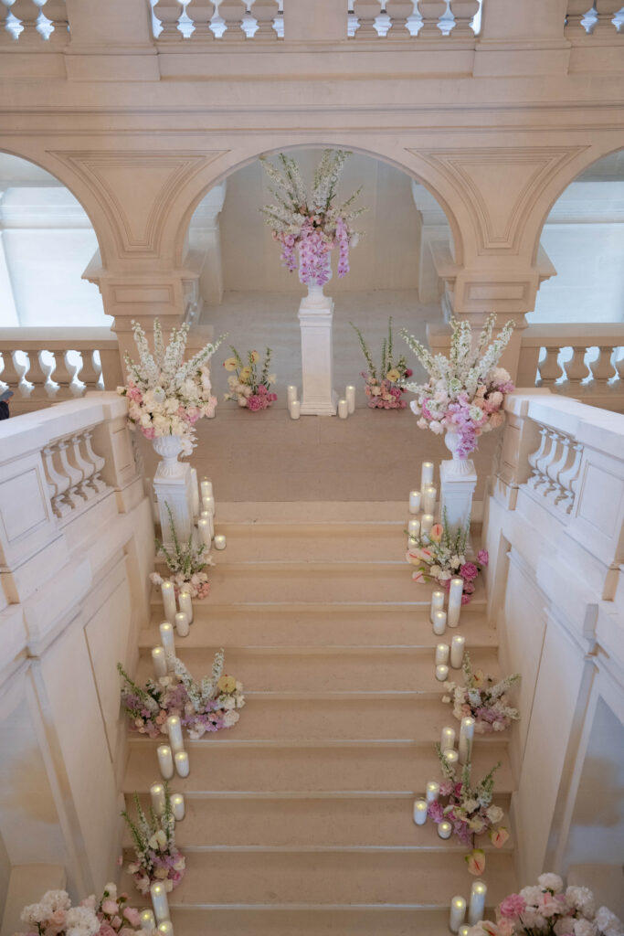 Eine große Treppe, geschmückt mit pastellfarbenen Blumen, weißen Kerzen und eleganten Dekorationen für eine Veranstaltung – eine Atmosphäre, die an einen „Automatisch gespeicherten Entwurf“ erinnert, bei dem jedes Detail mühelos raffiniert ist.