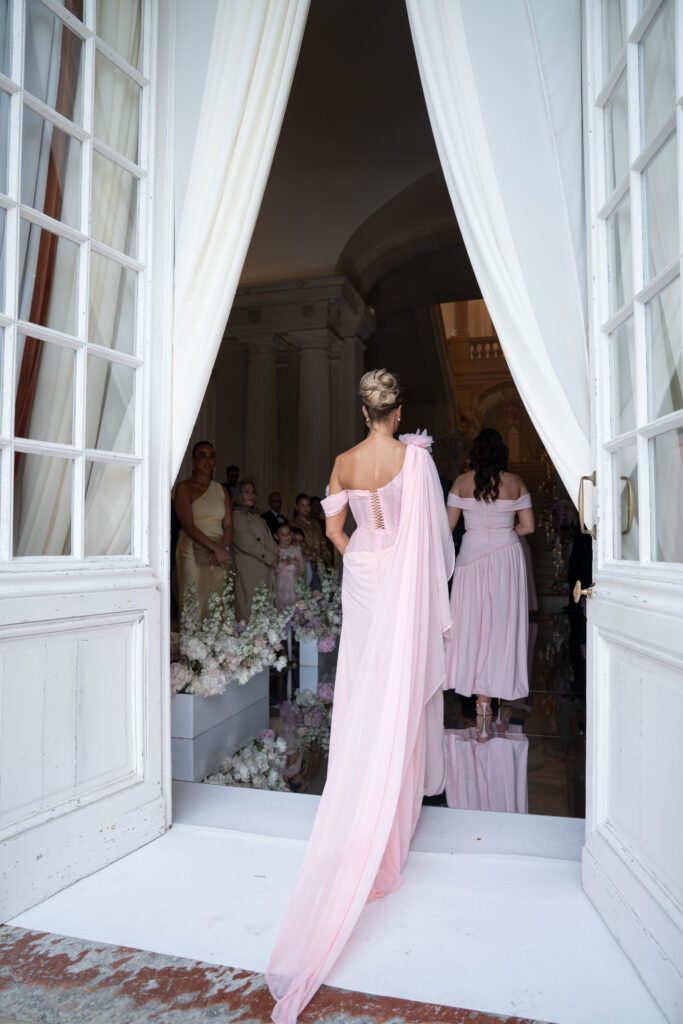 Eine Frau in einem rosa Kleid schreitet durch weiße Vorhänge in einen Raum voller Menschen und Blumen – von hinten gesehen – wie ein automatisch gespeicherter Entwurf eines eleganten Augenblicks, der in der Zeit festgehalten wurde.