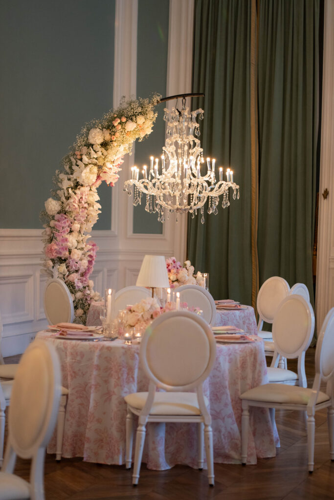 Ein elegantes Tischarrangement mit weißen Stühlen, einem Blumenbogen und einem Kronleuchter in einem sanft beleuchteten, edlen Raum – wie ein automatisch gespeicherter Entwurf einer raffinierten Veranstaltungsdekoration.