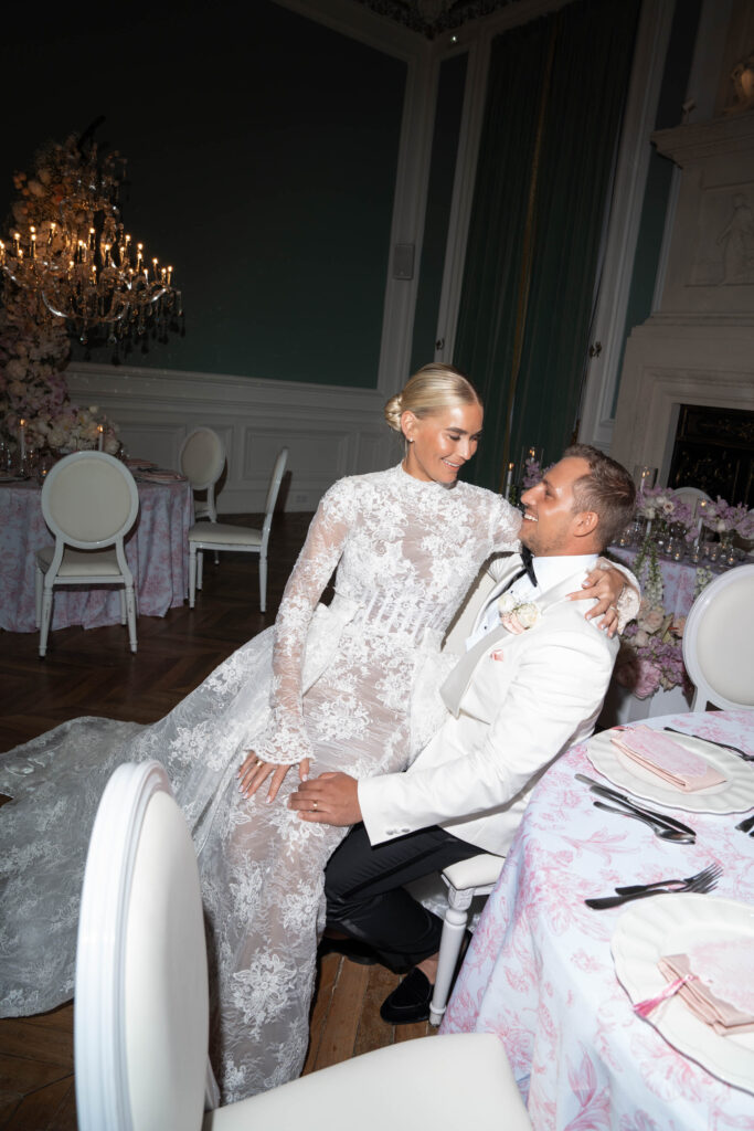 Bei einem eleganten Hochzeitsempfang bei Kerzenschein sitzt eine Braut in einem Spitzenkleid auf dem Schoß ihres Bräutigams; ihr Glück spiegelt die Magie des Abends wider wie ein automatisch gespeicherter Entwurf kostbarer Erinnerungen.