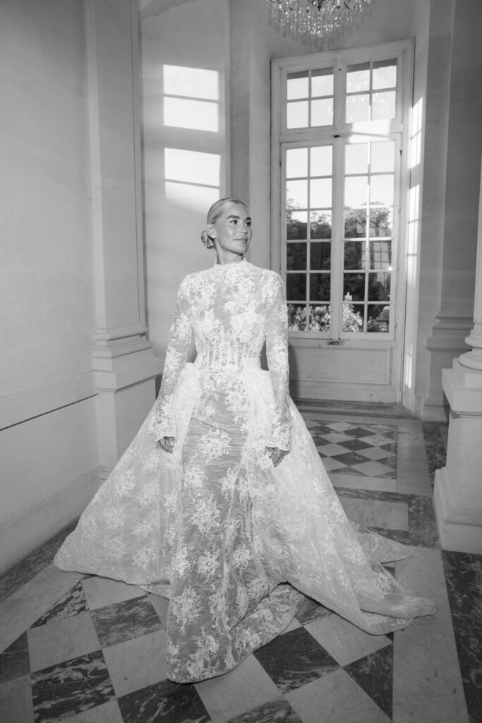 Eine Frau in einem langen Brautkleid aus Spitze steht in einem hellen, eleganten Raum mit hohen Fenstern, als wäre sie das Motiv eines „automatisch gespeicherten Entwurfs“ – eines mühelos schönen Entwurfs, eingefangen in zeitlosem Licht.