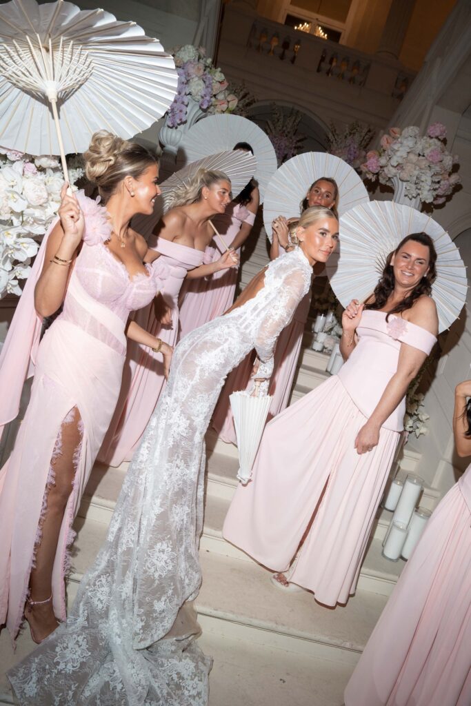 Eine Braut in einem Spitzenkleid posiert auf einer Treppe zusammen mit ihren Brautjungfern in rosa Kleidern; alle halten weiße Sonnenschirme in der Hand und fangen so einen eleganten Moment ein, der an einen automatisch gespeicherten Entwurf erinnert.