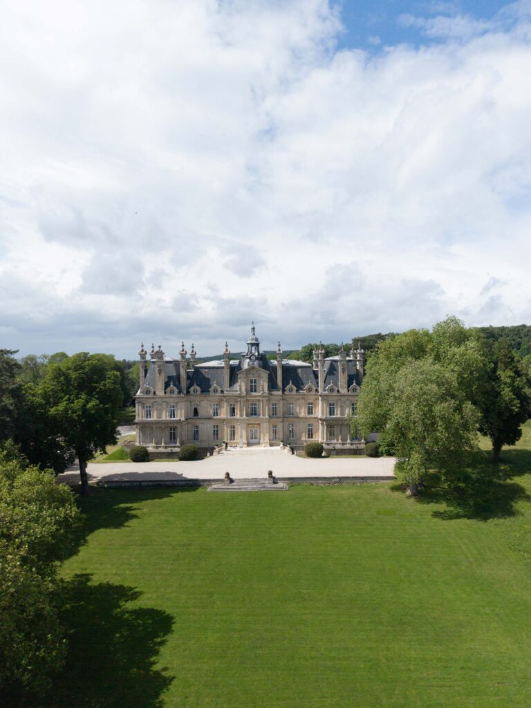 Ein prächtiges Schloss mit kunstvollen Details, umgeben von Bäumen und einer großen grünen Rasenfläche unter einem teilweise bewölkten Himmel, verkörpert die zeitlose Eleganz eines automatisch gespeicherten Entwurfs, der in atemberaubender Architektur zum Leben erweckt wurde.