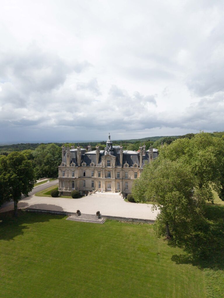 Ein prächtiges Schloss aus Stein mit einem kunstvoll verzierten Dach steht inmitten grüner Rasenflächen und Bäume unter einem bewölkten Himmel und erinnert an die Eleganz eines automatisch gespeicherten Entwurfs, der zum Leben erweckt wurde.
