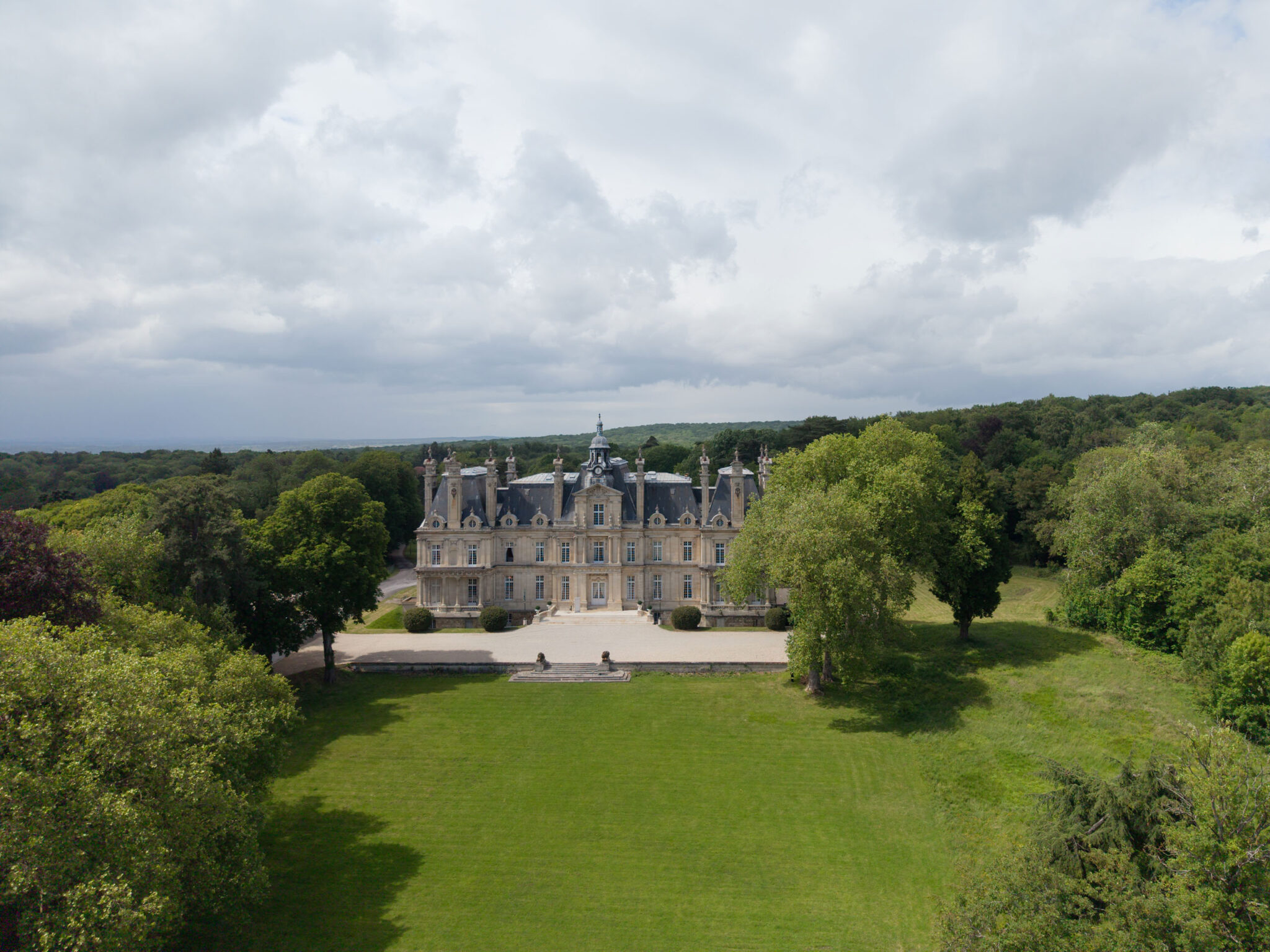 Ein prächtiges Schloss aus Stein mit Türmen, umgeben von Bäumen und einer großen Rasenfläche unter einem bewölkten Himmel, steht da wie ein „automatisch gespeicherter Entwurf“ – ein Entwurf von zeitloser Eleganz, eingebettet in die Natur.