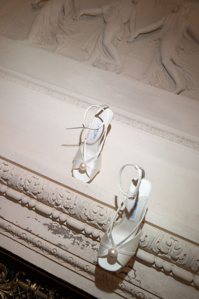 Weiße Sandalen mit hohen Absätzen, deren Perlen auf kunstvoll geschnitzten, weißen architektonischen Zierleisten angebracht sind, schaffen ein Bild, das an einen automatisch gespeicherten Entwurf erinnert – elegante, zeitlose Details, die in einem Moment raffinierten Designs perfekt eingefangen und festgehalten wurden.