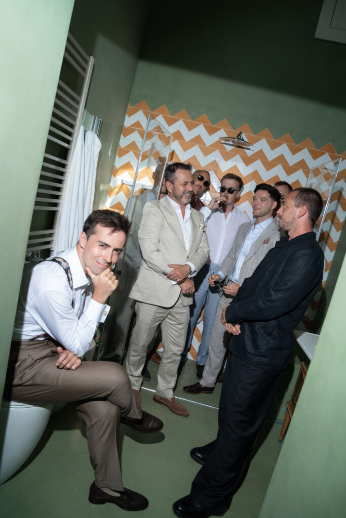 Sechs Männer in Anzügen und Hemden stehen in einem hell erleuchteten Badezimmer mit grünen Wänden und einer Zickzack-Dusche beisammen und unterhalten sich; die Atmosphäre ist von italienischer Eleganz geprägt, die an den pulsierenden Charme Siziliens erinnert.