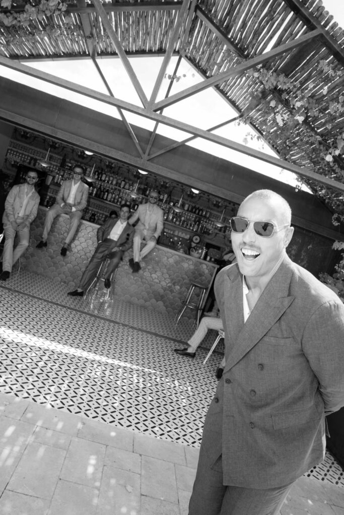 Ein Mann mit Sonnenbrille lächelt vor vier sitzenden Männern in einer Außenbar mit gemustertem Fliesenboden und fängt so die entspannte Atmosphäre Siziliens, Italien, ein.