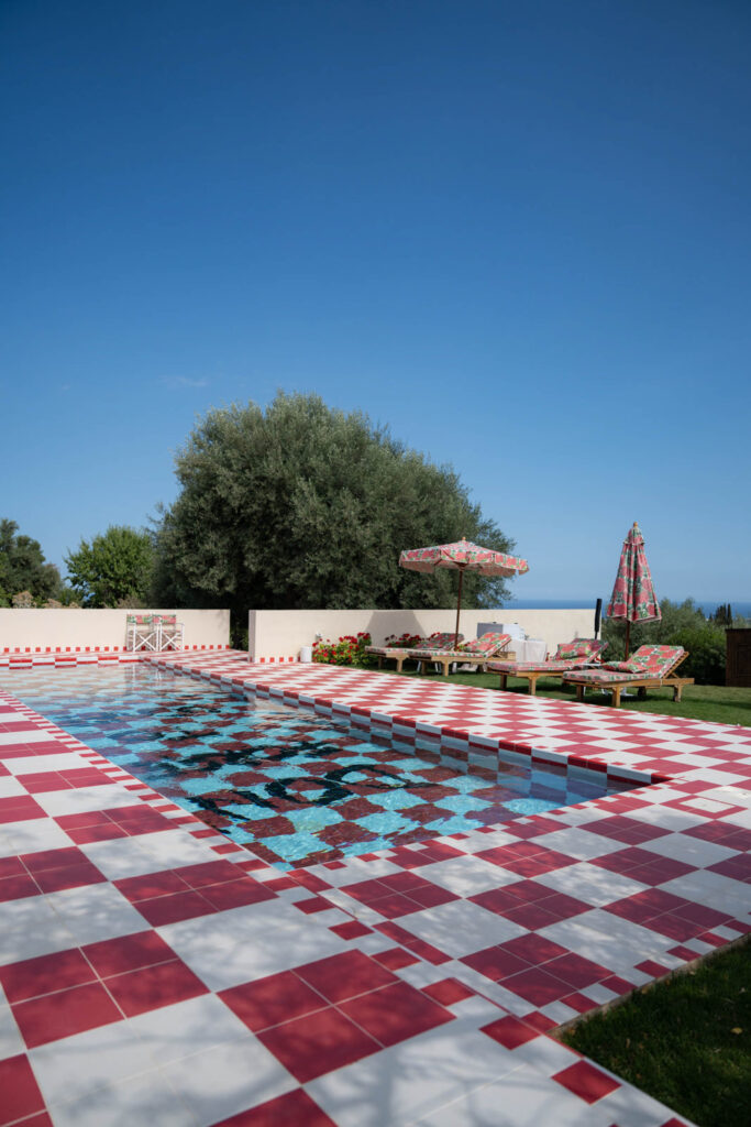 Eine rot-weiß karierte Poolterrasse mit einem gemusterten Pool, Sonnenliegen, Sonnenschirmen, Bäumen und strahlend blauem Himmel – eine Atmosphäre, die sich anfühlt wie ein automatisch gespeicherter Entwurf von Urlaubsentspannung, der hier lebendig zum Leben erweckt wurde.