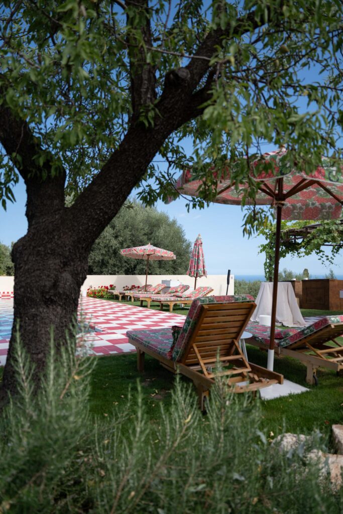 Holzliegestühle und rote Sonnenschirme am Pool, unter einem Baum, umgeben von üppigem Grün und blauem Himmel – das alles versprüht den Charme Siziliens, Italien.