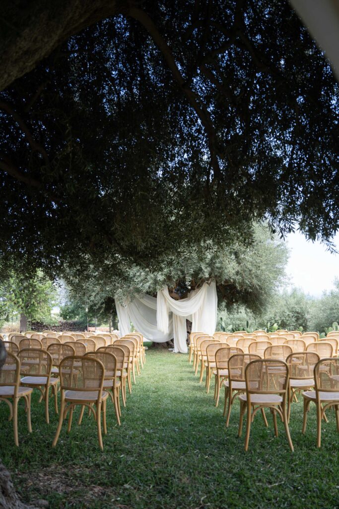 Unter einem großen Baum stehen Stuhlreihen vor einem mit weißen Tüchern drapierten Bogen – alles für eine Hochzeitszeremonie im Freien in der romantischen Landschaft Siziliens, Italien.