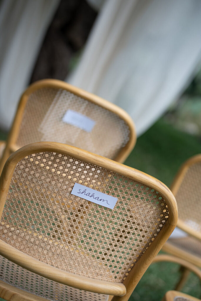 Nahaufnahme von Holzstühlen mit geflochtenen Rückenlehnen, die an traditionelle italienische Handwerkskunst erinnern; einer davon trägt mit einem Namensschild aus Papier die Aufschrift „Shadam“ – und versprüht den entspannten Charme Siziliens.