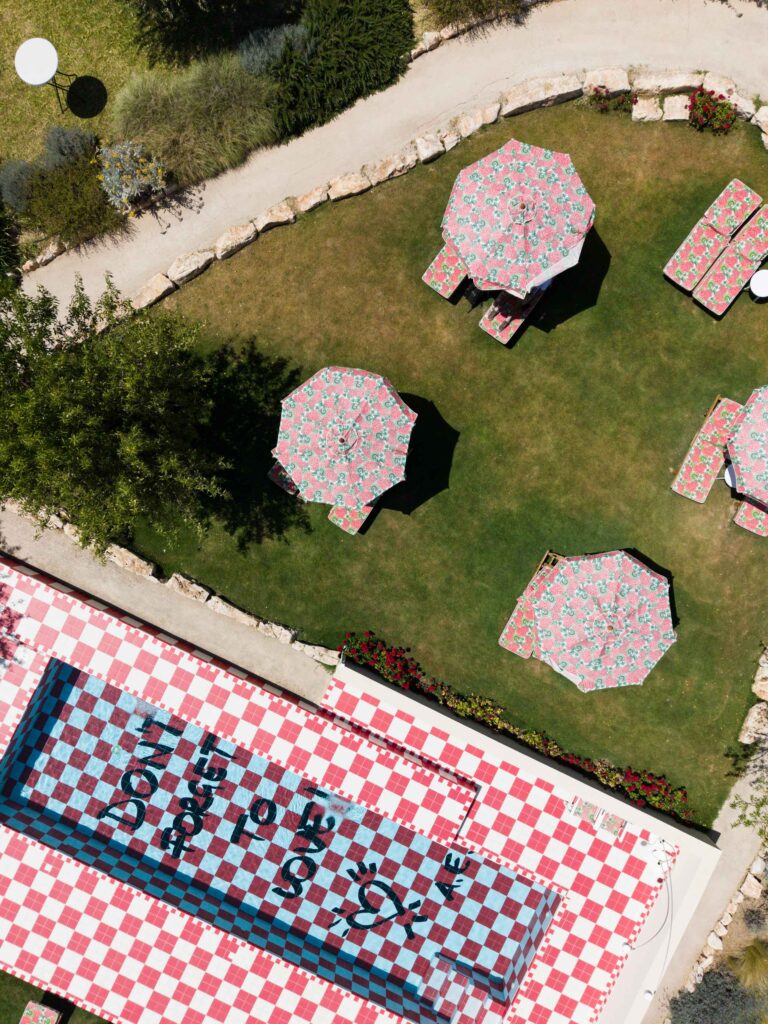 Luftaufnahme eines mit rotem Karomuster verzierten Pools, Sonnenschirmen und Picknicktischen auf einer Rasenfläche mit einem Steinweg, die den entspannten Charme des sonnigen Siziliens in Italien einfängt.
