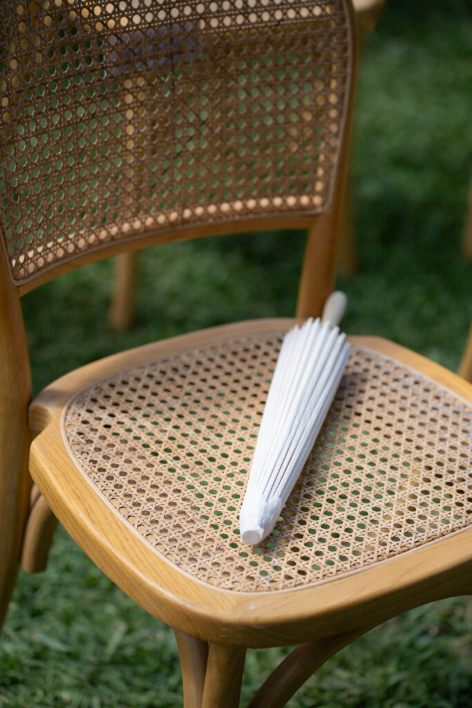 Ein zusammengefalteter weißer Papiersonnenschirm liegt auf einem geflochtenen Rattanstuhl mit Holzgestell im Freien auf dem Rasen und erinnert an die entspannte Eleganz Siziliens in Italien.