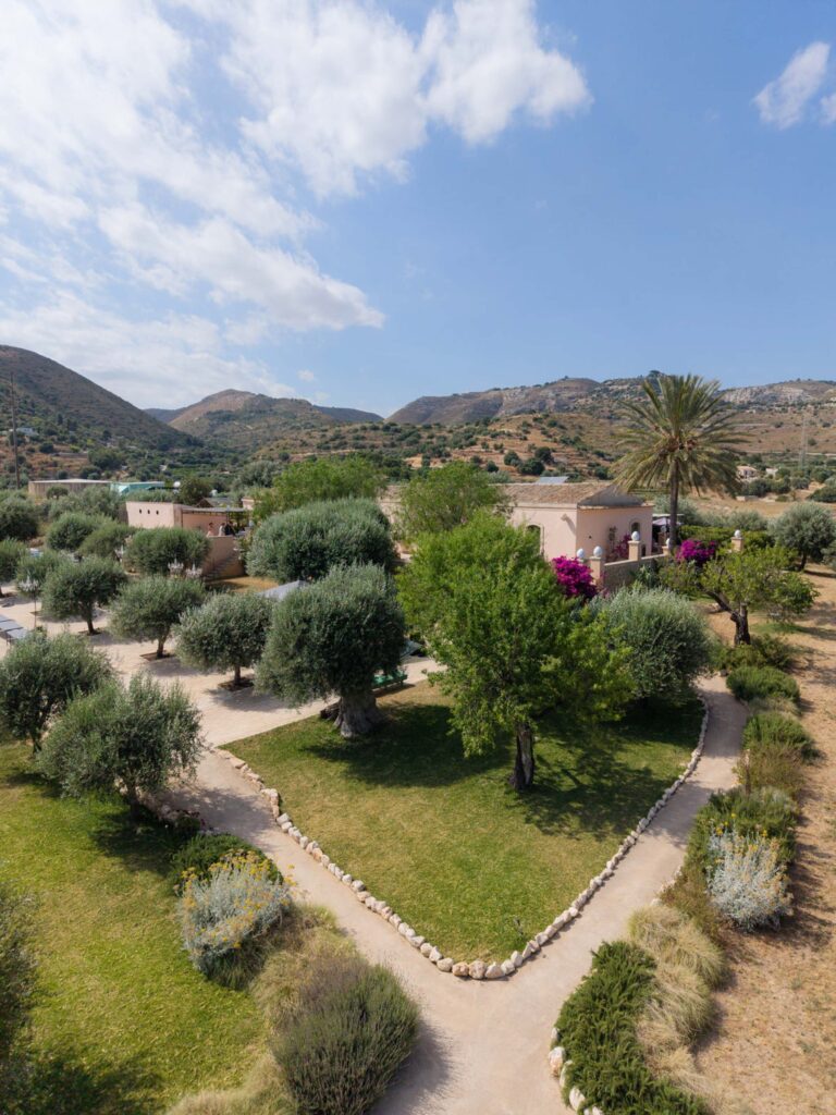 Ein mediterraner Garten mit Olivenbäumen, Wegen und Gebäuden vor dem Hintergrund von Hügeln unter einem teilweise bewölkten Himmel versetzt den Betrachter in den ruhigen Charme Siziliens in Italien.
