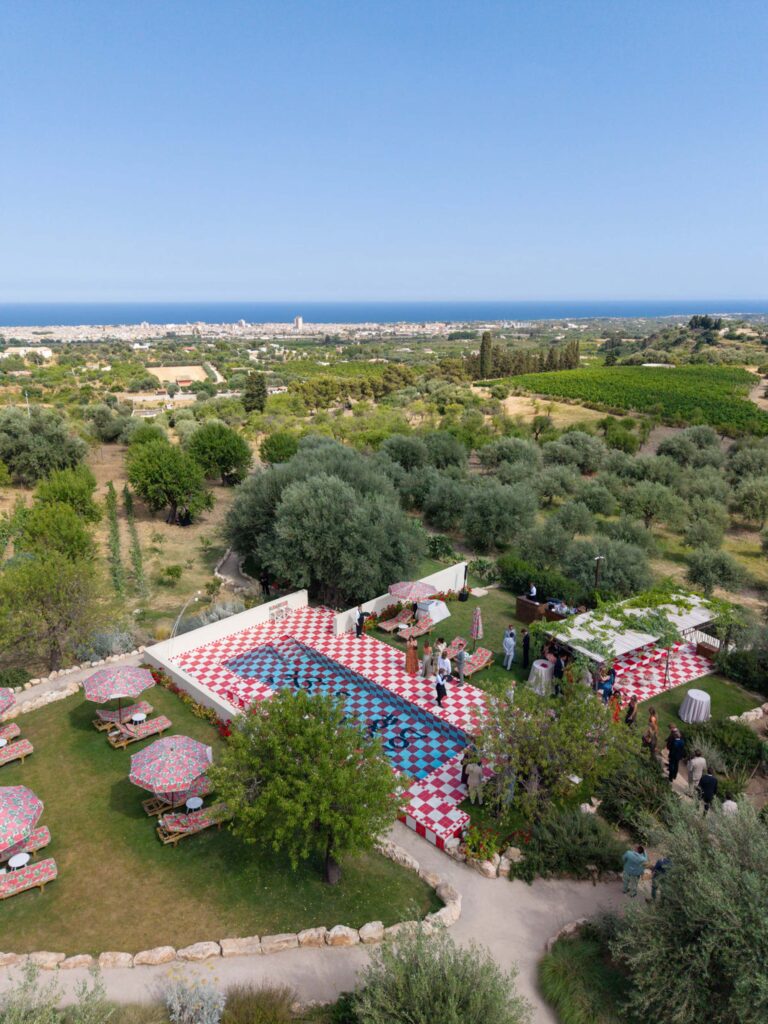 Luftaufnahme einer Veranstaltung im Freien mit einem rot-weiß karierten Boden, umgeben von Bäumen und mit Blick auf das in der Ferne liegende Meer, die den lebendigen Charme Siziliens, Italien, einfängt.