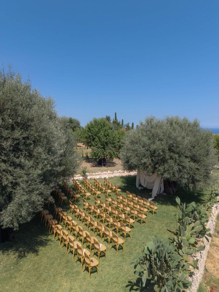 An einem sonnigen Tag stehen im Freien unter Bäumen Reihen von Holzstühlen, die auf ein mit weißem Stoff drapiertes Festzelt ausgerichtet sind und die entspannte Eleganz einer Hochzeit in Sizilien, Italien, heraufbeschwören.