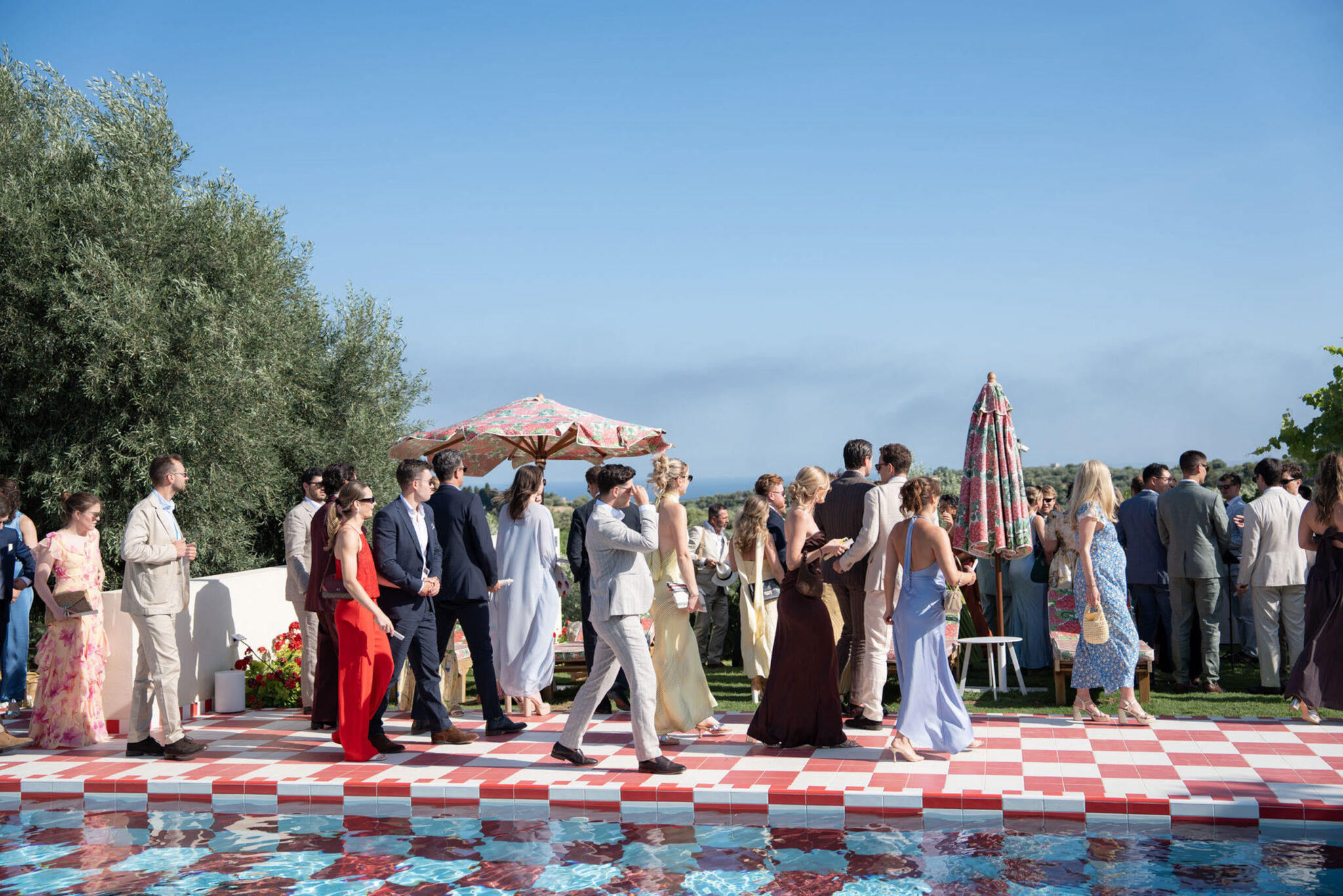 Menschen in festlicher Kleidung versammeln sich unter blauem Himmel an einem Pool mit rot kariertem Terrassenboden und unterhalten sich bei einer Veranstaltung im Freien, die den pulsierenden Geist Italiens widerspiegelt und an stilvolle Zusammenkünfte in Sizilien erinnert.