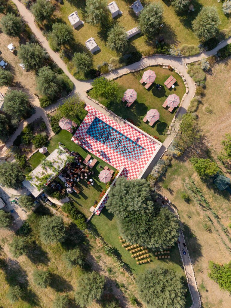 Luftaufnahme einer Hochzeit in Sizilien mit einer rot-weiß karierten Tanzfläche und Sitzgelegenheiten im Freien unter Sonnenschirmen, die eine bezaubernde italienische Atmosphäre schaffen.