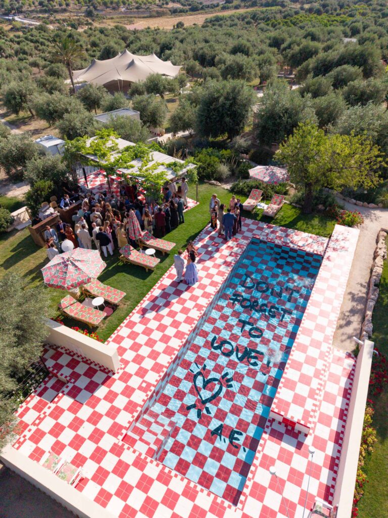 Eine Gruppe versammelt sich an einem Pool in Sizilien, auf dessen Boden die Worte „Vergiss nicht, mich zu lieben!“ zu lesen sind, umgeben von üppigem italienischem Grün.