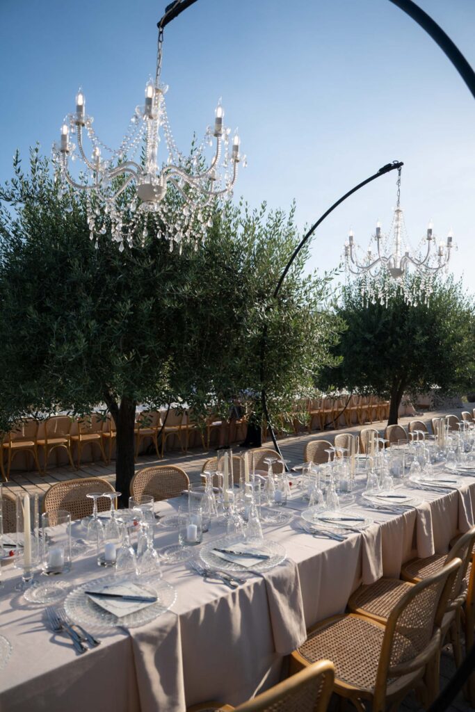 Ein eleganter Esstisch im Freien unter Kronleuchtern, umgeben von Bäumen, gedeckt mit Glasgeschirr und Tischwäsche in neutralen Farbtönen, versetzt den Gast in den zeitlosen Charme des italienischen Stils, inspiriert von den bezaubernden Landschaften Siziliens.