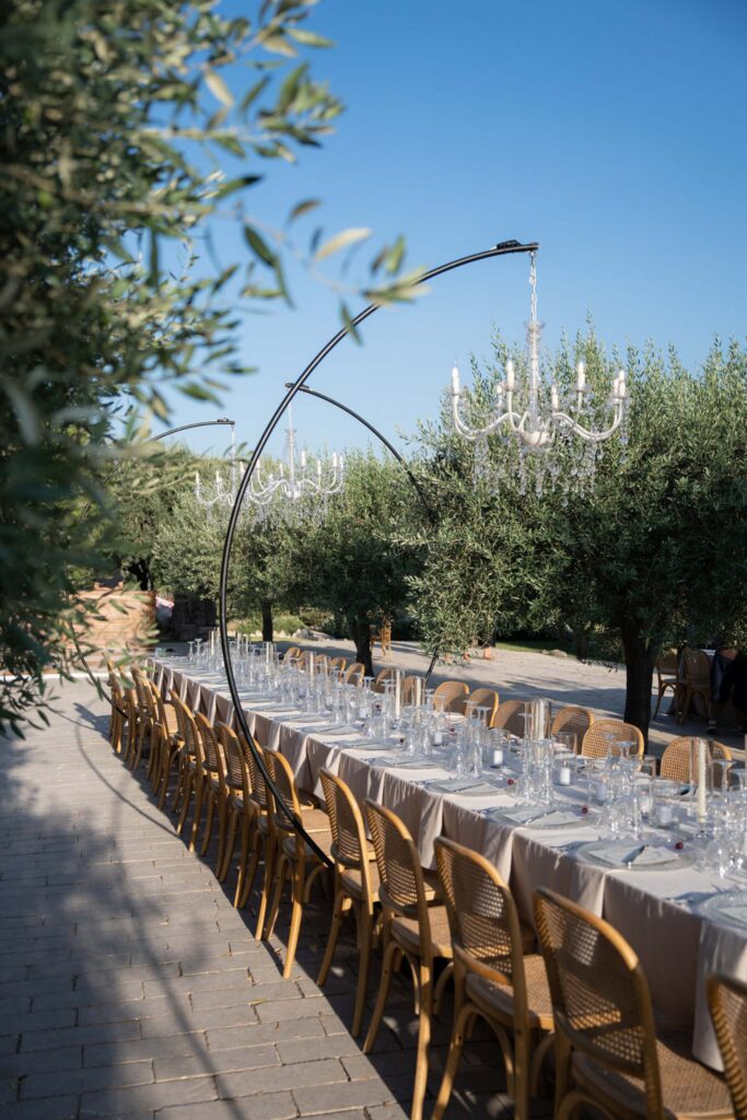 Ein langer Tisch, gedeckt für ein Essen im Freien unter Kronleuchtern, umgeben von Bäumen und Holzstühlen auf einem Steinweg, der den rustikalen Charme Siziliens in Italien heraufbeschwört.