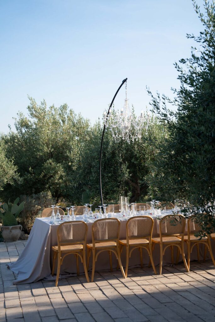 Ein eleganter Esstisch für den Außenbereich, der an italienischen Charme erinnert, mit Holzstühlen, einer grauen Tischdecke, edlem Glasgeschirr und einem hängenden Kronleuchter – perfekt für einen Hauch sizilianischer Raffinesse.