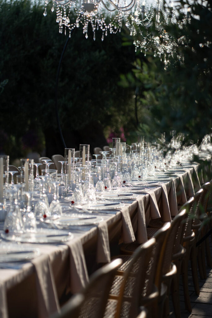 Ein langer Tisch im Freien, gedeckt mit Gläsern und weißen Tischdecken, unter einem Kronleuchter in sanftem Licht, vermittelt die zeitlose Eleganz Siziliens, Italien.