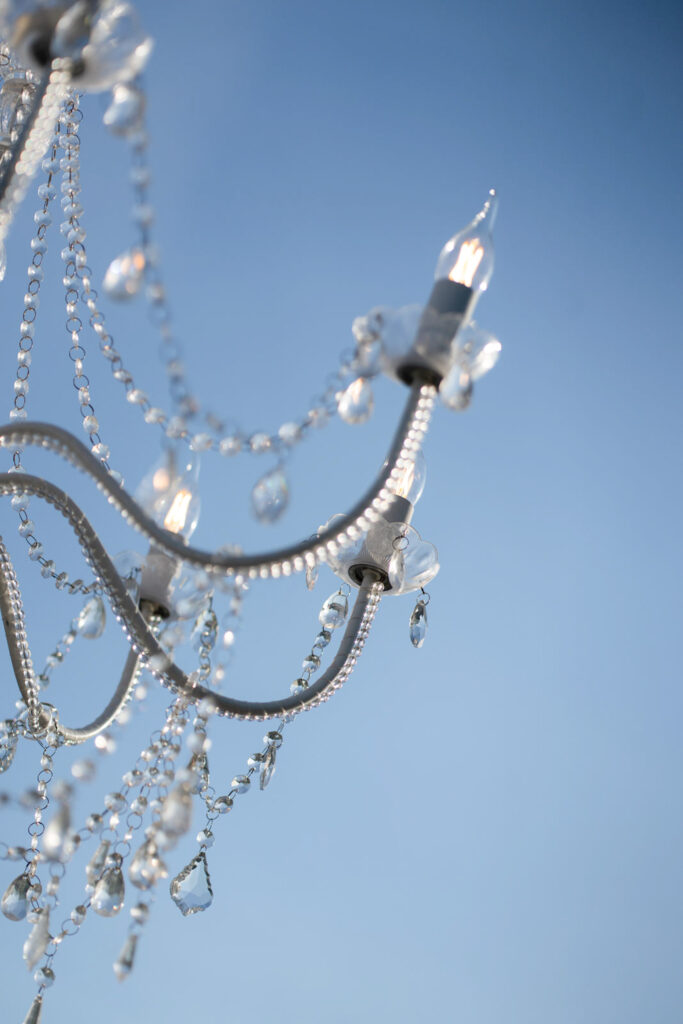 Ein Kristallleuchter mit brennenden Glühbirnen und herabhängenden Perlen vor einem strahlend blauen Himmel, der die Eleganz des italienischen Designs und den sonnendurchfluteten Charme Siziliens heraufbeschwört.
