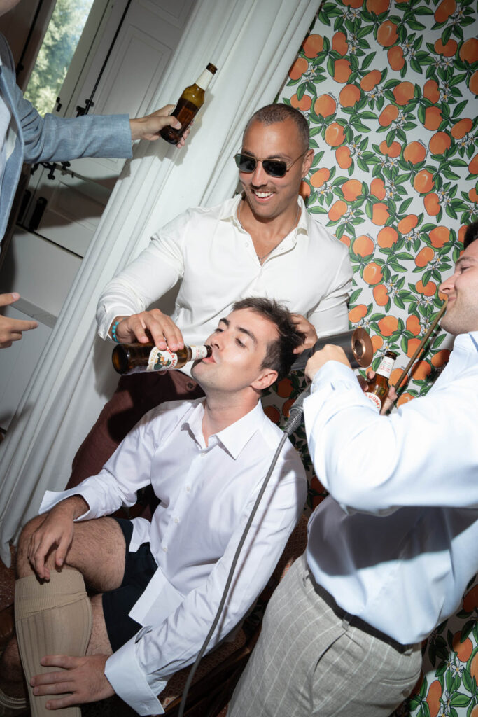 Ein Mann mit Sonnenbrille grinst, während ein anderer Bier trinkt; eine lebhafte Szene, die an Italien erinnert, in einem hellen Raum mit orangefarbener Tapete.