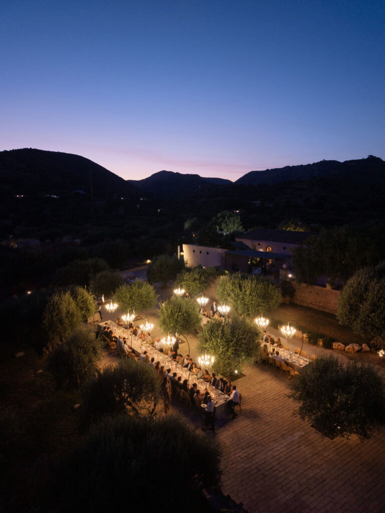 Eine Dinnerparty im Freien bei Einbruch der Dämmerung in Sizilien, Italien, bei der Kronleuchter einen langen Tisch beleuchten, der von Bäumen und Hügeln umgeben ist.