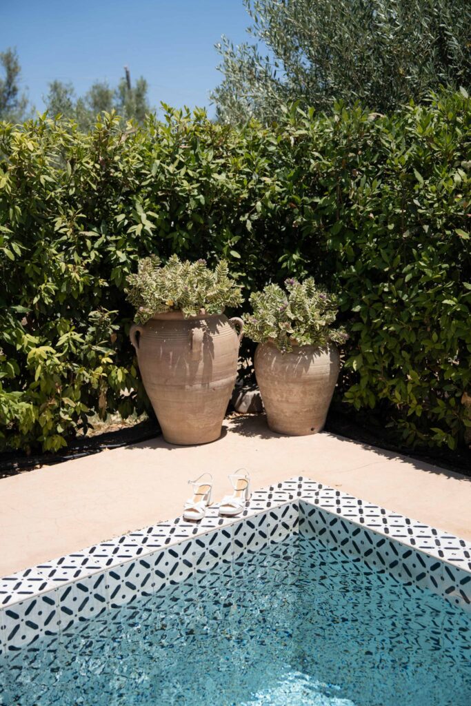 Der Rand eines mit Fliesen ausgelegten Pools mit zwei großen Pflanztöpfen und Sandalen auf dem Boden, umgeben von grünen Sträuchern, die den ruhigen Charme Siziliens in Italien heraufbeschwören.