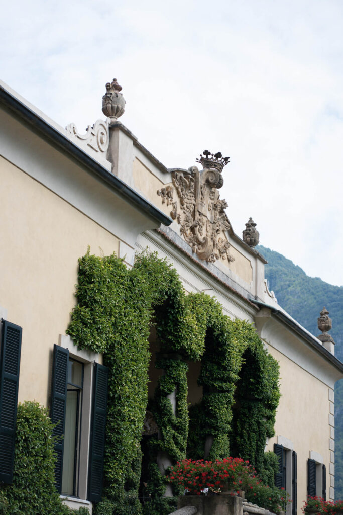 Ein mit Efeu bewachsenes Gebäude mit kunstvollen Steinverzierungen, schwarzen Fensterläden und roten Blumen vor einer Bergkulisse – eine fast malerische Szene, die an einen automatisch gespeicherten Entwurf mit klassischem europäischem Charme erinnert.