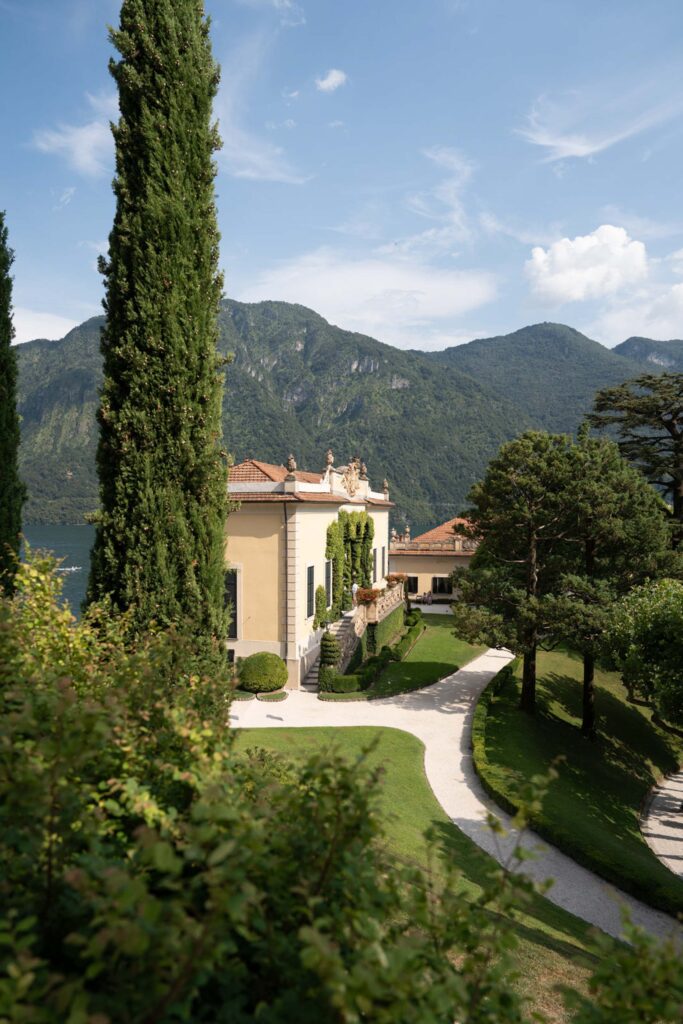Eine gelbe Villa am See, umgeben von Bäumen, mit einem gewundenen Weg und grünen Bergen im Hintergrund unter blauem Himmel, gleicht einem automatisch gespeicherten Entwurf von stiller Schönheit.