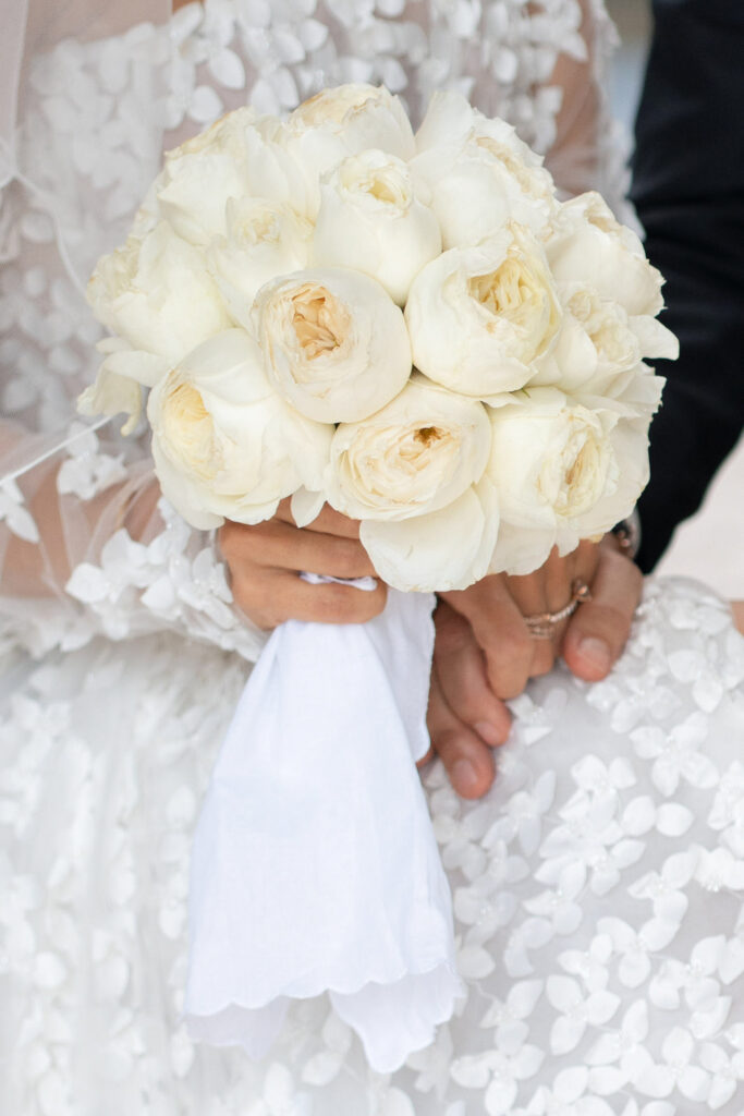 Eine Braut, die einen Strauß weißer Rosen hält, ein Kleid aus Blumenspitze trägt und mit einer anderen Person Händchen hält – ein Moment, der so zeitlos und elegant ist wie ein automatisch gespeicherter Entwurf.