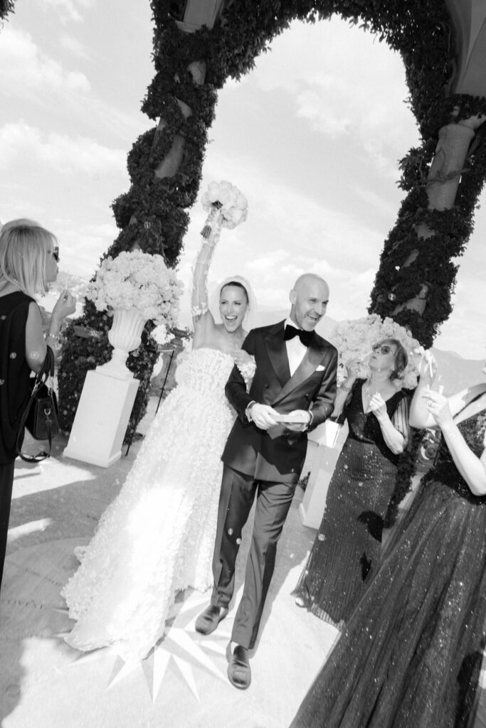 Ein Brautpaar lächelt, während es Arm in Arm unter einem Torbogen hindurchgeht, während die Gäste auf ihrer Hochzeit feiern – ein Moment, der sich wie ein automatisch gespeicherter Entwurf puren Glücks anfühlt.