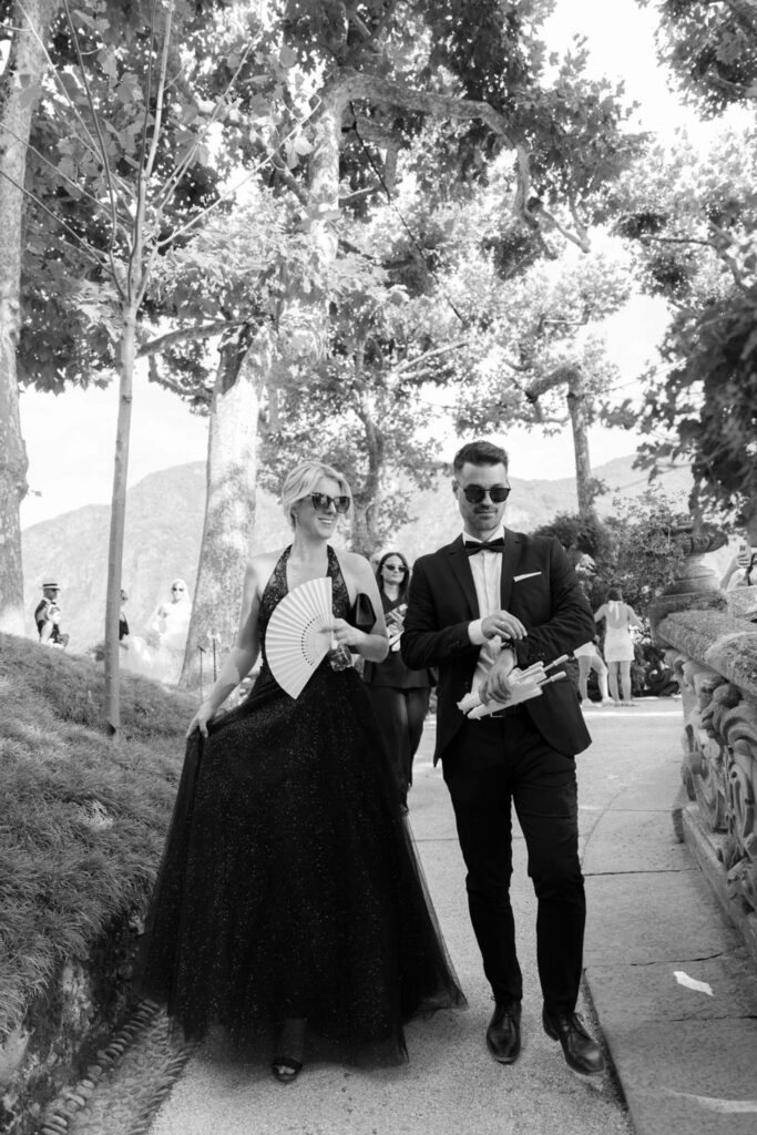 Zwei Personen in eleganter Kleidung spazieren draußen zwischen den Bäumen in der Nähe des Comer Sees; eine von ihnen hält einen Fächer in der Hand, beide tragen eine Sonnenbrille und strahlen eine elegante italienische Atmosphäre aus.