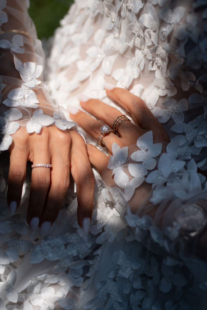 Nahaufnahme von Händen in einem Kleid aus Blumenspitzen, an denen zahlreiche Ringe stecken, während Licht und Schatten über sie hinwegtanzen – was an die Eleganz des Comer Sees in Italien erinnert.