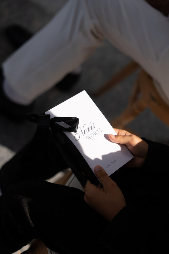 Eine Person, die ein weißes Büchlein mit dem Titel „Noah’s VOWS“ auf dem Einband in der Hand hält, das mit einem schwarzen Band zusammengebunden ist, vor der malerischen Kulisse des Comer Sees in Italien.