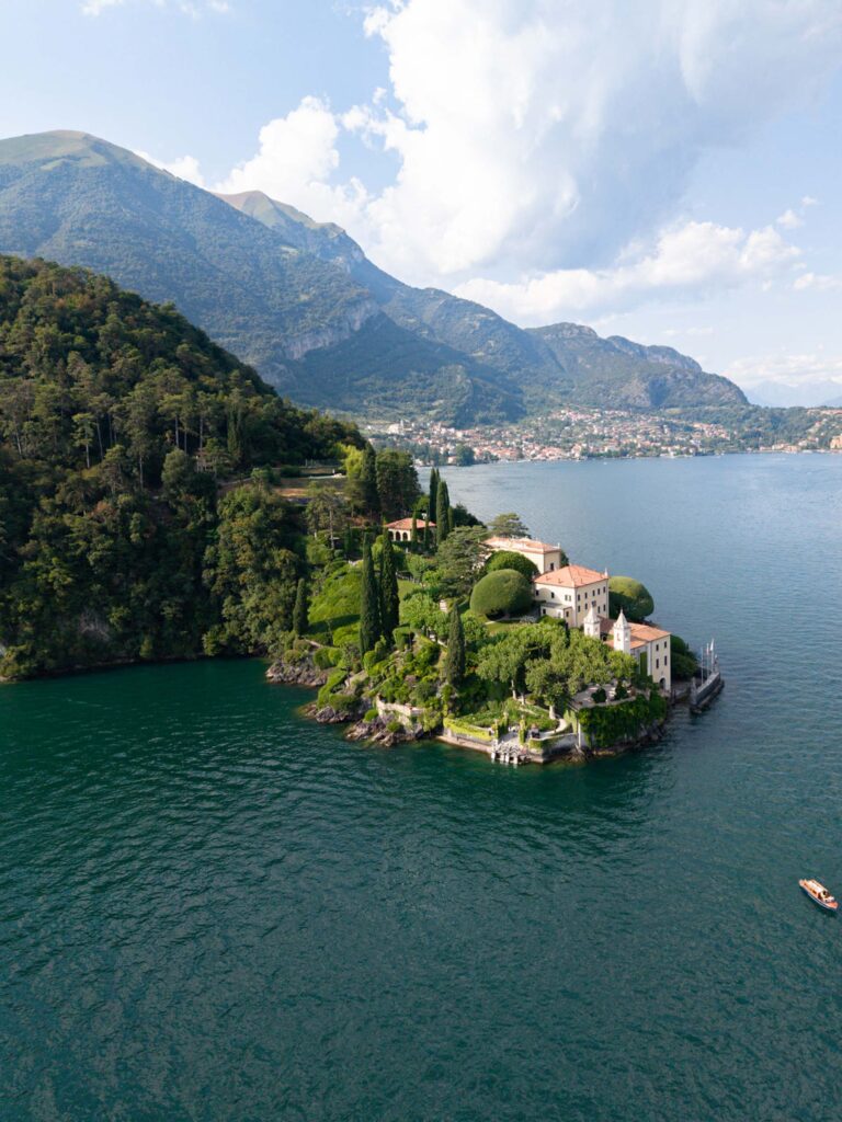 Eine von Bäumen umgebene Villa auf einer kleinen Halbinsel, die in einen See hineinragt, mit Bergen im Hintergrund, bietet einen atemberaubenden Blick auf den Comer See in Italien.